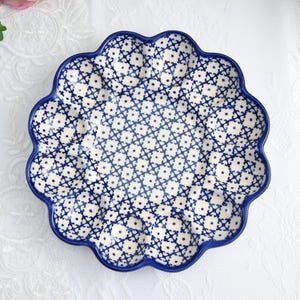 Può includere: Un piatto in ceramica con bordo smerlato, con un motivo floreale blu e bianco. Il piatto ha un bordo blu scuro e uno sfondo color crema. Il design è un motivo ripetitivo di piccoli fiori stilizzati.