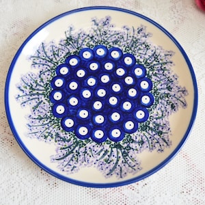 May include: A white ceramic plate with a blue and purple floral design. The centre of the plate features a cluster of blue circles with white centres, surrounded by purple flowers with green stems.
