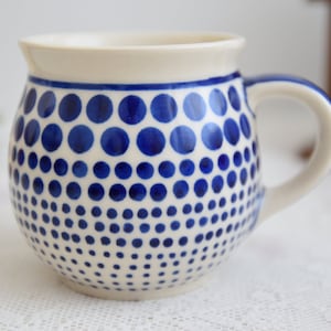 Könnte beinhalten: Eine weiße Keramiktasse mit einem blauen Tupfenmuster. Die Tasse hat einen abgerundeten Körper und einen Henkel.