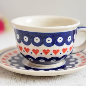 May include: A white ceramic teacup and saucer set with a blue and red heart pattern. The cup has a white interior and a rounded handle.