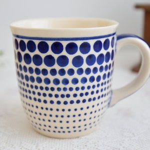 Può includere: Una tazza in ceramica bianca con un motivo a pois blu. I pois sono disposti in un motivo graduato, con pois più grandi in alto e pois più piccoli in basso.