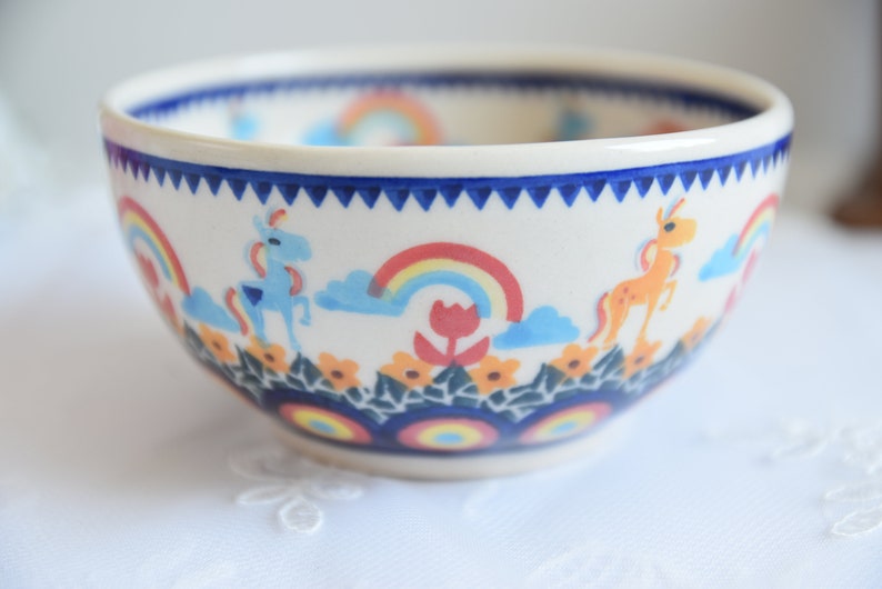 Op de afbeelding: Een witte keramische kom met een blauwe en witte rand en een kleurrijk ontwerp van regenbogen, bloemen en eenhoorns.
