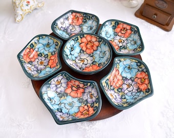 May include: A set of seven ceramic serving dishes with a floral design in shades of blue, pink, and orange. The dishes are arranged in a circle on a wooden base.
