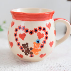 Könnte beinhalten: Eine weiße Keramiktasse mit rotem Rand und Henkel. Die Tasse ist mit einem herzförmigen Design verziert, das rote Herzen, blaue Blumen und drei Teddybären in Braun, Orange und Blau zeigt.