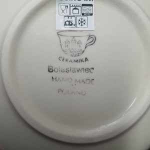 May include: Off-white ceramic plate with a stamped design and text. The plate features a cup illustration with castle-like structures and the words "CERAMIKA Bolesławiec HAND MADE POLAND". A sticker indicates the plate is microwave, dishwasher, oven, and freezer safe.