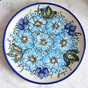 May include: A white ceramic plate with a blue and white floral design. The plate features a bouquet of blue flowers with yellow centres and green leaves.