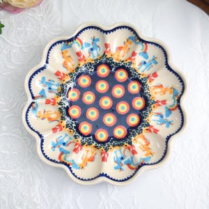 Peut inclure: Assiette à œufs en céramique avec un bord festonné. L'assiette présente une base crème avec un motif de licorne bleu, orange et jaune. Le centre de l'assiette est bleu foncé avec des cercles jaunes et oranges. Le bord est orné d'un bord festonné bleu foncé.