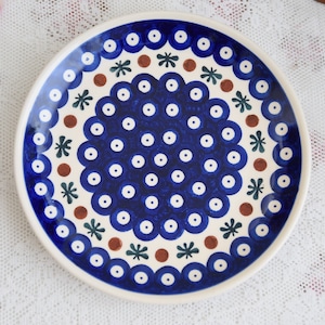 Può includere: Un piatto in ceramica blu e bianco con un motivo decorativo di cerchi, fiori e punti. Il piatto ha uno sfondo bianco con accenti blu e marroni.