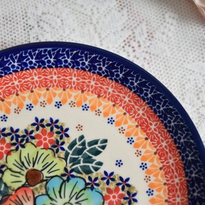 May include: A white ceramic plate with a blue rim and a colorful floral design. The plate features a border of orange, red, and blue floral patterns.