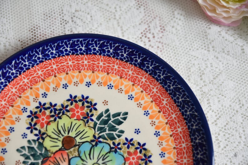 May include: A white ceramic plate with a blue rim and a colorful floral design. The plate features a border of orange, red, and blue floral patterns.