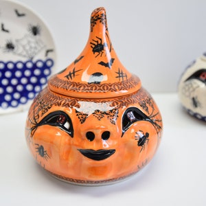 May include: Orange ceramic pumpkin-shaped container with a black and white spiderweb design and a black and white spider on the lid. The container has a black and white face with eyes, a nose, and a mouth.