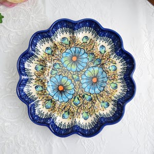 Può includere: Un piatto rotondo in ceramica con bordo smerlato e design floreale blu. Il piatto presenta tre grandi fiori blu con centri arancioni e accenti verdi, circondati da elementi floreali più piccoli su sfondo crema. Il bordo è blu scuro.