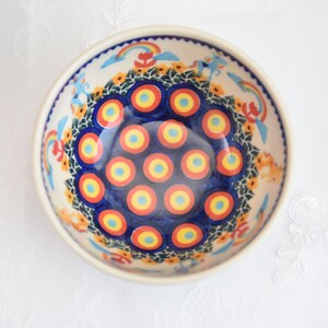 Op de afbeelding: Een blauwe en witte keramische kom met een kleurrijk ontwerp van concentrische cirkels in rood, geel en blauw. De kom heeft een witte rand en een bloemenpatroon rond de rand.