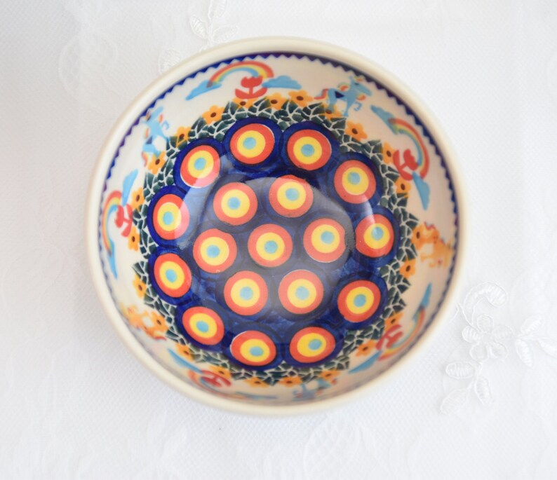 Op de afbeelding: Een blauwe en witte keramische kom met een kleurrijk ontwerp van concentrische cirkels in rood, geel en blauw. De kom heeft een witte rand en een bloemenpatroon rond de rand.