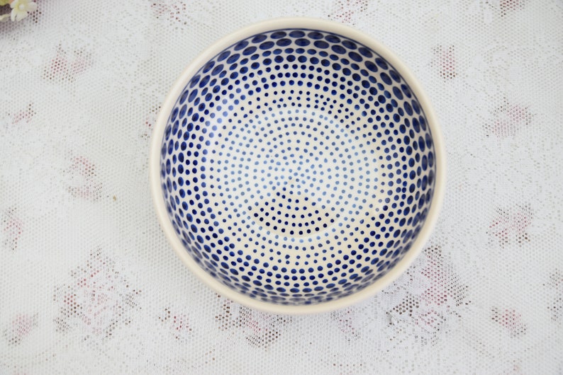 May include: A white ceramic bowl with a blue polka dot pattern. The dots are arranged in a concentric circle pattern.