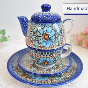 Peut inclure: Un ensemble théière et tasse en céramique bleu et blanc avec un motif floral. L'ensemble est fait à la main et comprend une théière, une tasse et une soucoupe. La théière a un couvercle et une poignée. La tasse a une poignée. La soucoupe est ronde. L'ensemble est sur une nappe en dentelle blanche.