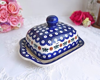 Könnte beinhalten: Blau-weiße Keramik-Butterdose mit passendem Deckel. Der Deckel ist mit einem Muster aus weißen Punkten und floralen Akzenten verziert. Die Dose steht auf einer weißen Spitzentischdecke.