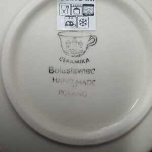 May include: White ceramic saucer with a black stamp that reads "Ceramika Boleslawiec Hand Made in Poland" and a symbol of a cup with a castle on it. The stamp also includes a label that says "Using in:" with symbols for microwave, oven, dishwasher, and freezer safe.
