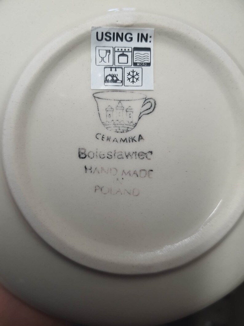 May include: White ceramic saucer with a black stamp that reads "Ceramika Boleslawiec Hand Made in Poland" and a symbol of a cup with a castle on it. The stamp also includes a label that says "Using in:" with symbols for microwave, oven, dishwasher, and freezer safe.