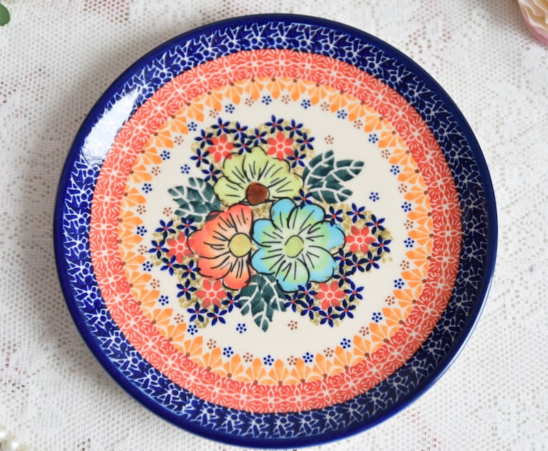 May include: A white ceramic plate with a blue and orange floral design. The plate features a central floral motif with pink, blue, and yellow flowers. The plate is surrounded by a blue and white border with intricate patterns.