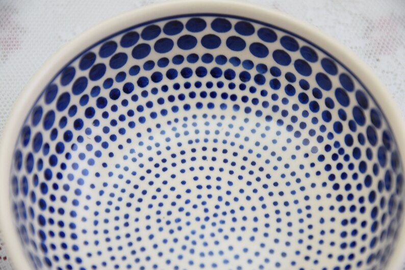 May include: A white ceramic bowl with a blue polka dot pattern. The dots are arranged in a graduated pattern, with larger dots at the top and smaller dots at the bottom.