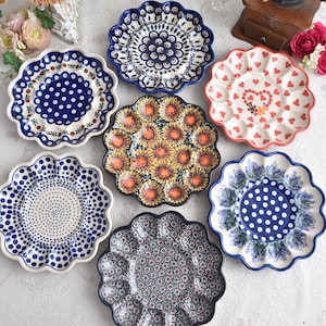 May include: A collection of decorative ceramic plates with scalloped edges. The plates feature various patterns, including polka dots, floral designs, and a sunflower motif. Colors include blue, white, red, and yellow.