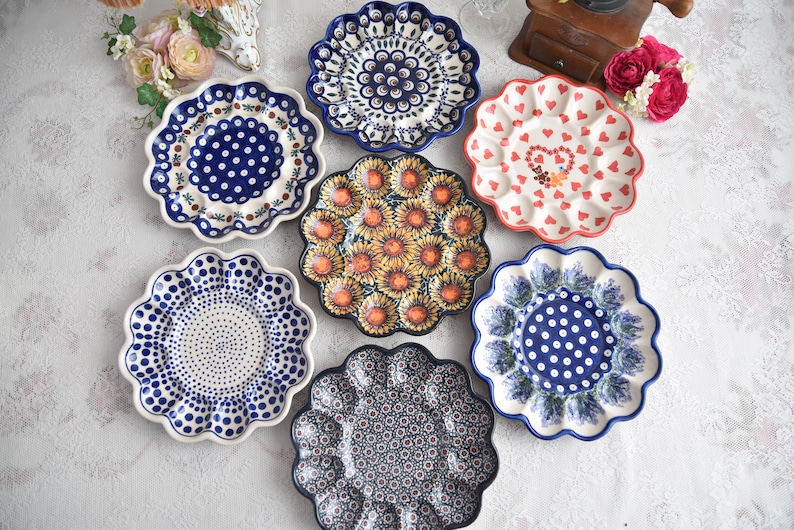 May include: A collection of decorative ceramic plates with scalloped edges. The plates feature various patterns, including polka dots, floral designs, and a sunflower motif. Colors include blue, white, red, and yellow.