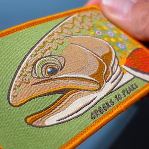May include: A rectangular embroidered patch featuring a detailed trout head in shades of tan, brown, and green. The fish has blue and red spots, and the text CREELS TO PEAKS is stitched in dark thread on a light green background with an orange border.