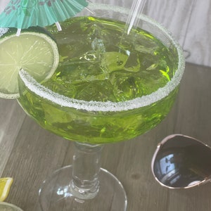 May include: A margarita cocktail in a stemmed glass, rimmed with salt. The drink is green with ice cubes, a lime slice, and a small green paper umbrella. Sunglasses are next to the glass.