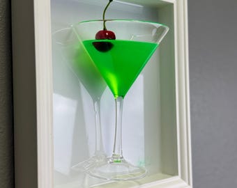 May include: A framed green cocktail with a cherry on top. The cocktail is in a martini glass and is framed in a white frame.