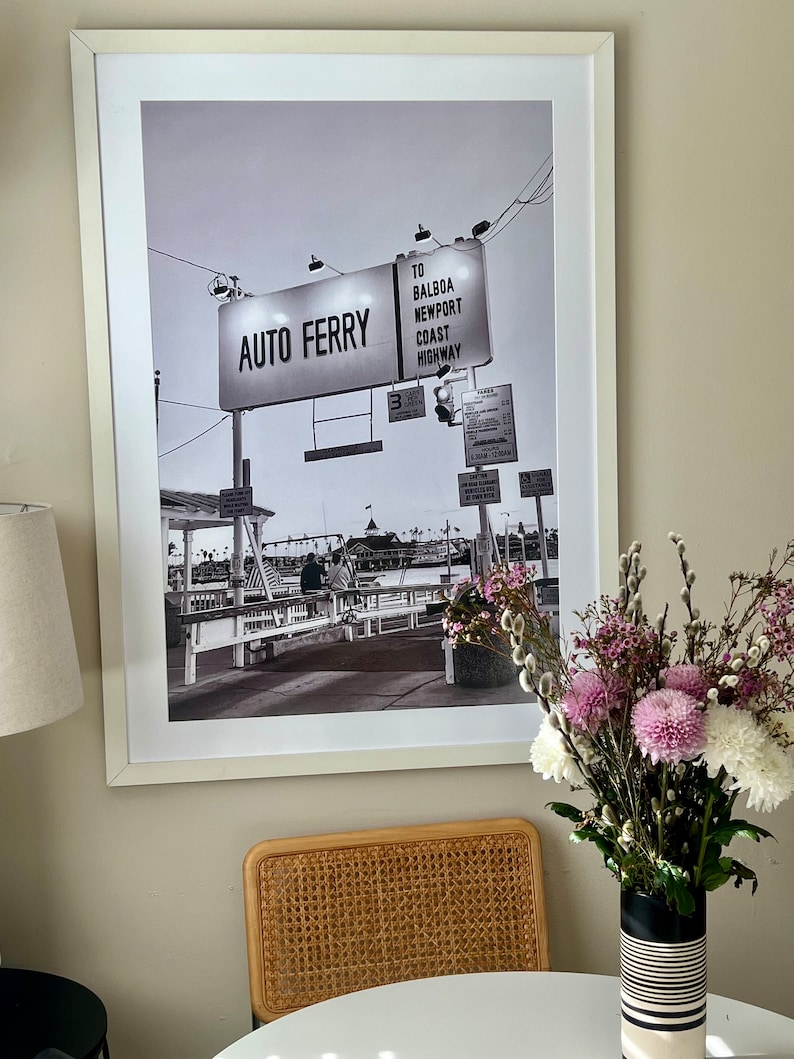 May include: A black and white photograph of a ferry terminal with a sign that reads "Auto Ferry" and "To Balboa Newport Coast Highway". The photo is framed in a white frame and hanging on a wall.
