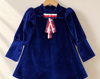 May include: A long-sleeved navy blue velvet dress featuring a high neckline and a decorative bow at the center chest. The bow is made of red, white, and navy blue striped ribbon with small gold-toned bee embroidery accents throughout.