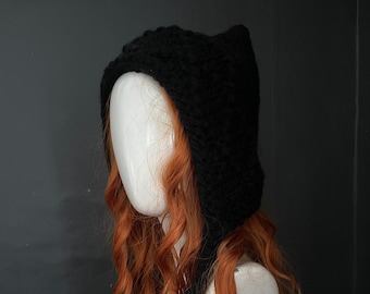 May include: Black knitted hooded hat with a long, flowing tail. The hat is worn on a mannequin with long, curly, auburn hair.