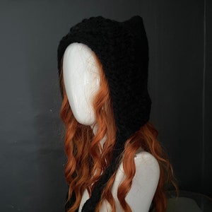 May include: Black knitted hooded hat with a long, flowing tail. The hat is worn on a mannequin with long, curly, auburn hair.