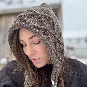 May include: A brown knitted hooded hat with a white and brown braided cord. The hat is made of a thick yarn and has a loose fit.