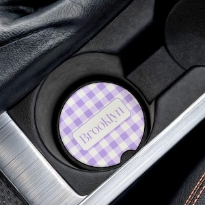May include: A car cup holder coaster with a purple and white gingham pattern. The coaster has the name "Brooklyn" printed on it in a white rectangular label. The coaster is placed inside a car's cup holder.