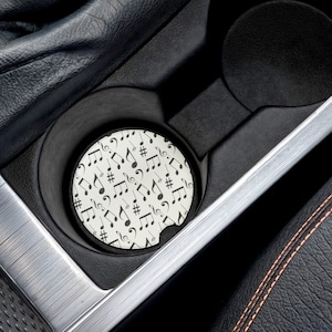 May include: A white round car coaster with a black musical note pattern. The coaster is in a car cup holder.