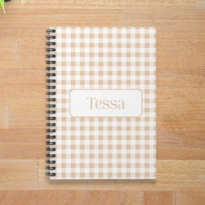 May include: A spiral-bound notebook with a tan and white gingham pattern. The notebook has the name "Tessa" on a rectangular label. A cup of coffee and pencils are on a wooden surface.