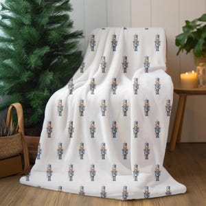 May include: A white plush blanket draped over a chair, featuring a repeating pattern of toy soldier illustrations. The soldiers are depicted in gray and blue uniforms with black boots and hats. The blanket is soft and cozy, perfect for the holiday season.