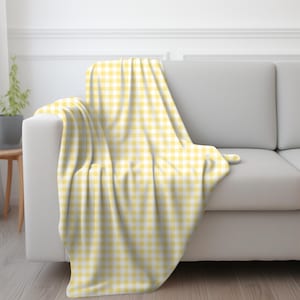 May include: A yellow and white gingham pattern throw blanket draped over a white sofa. The blanket's pattern is a classic checkered design, adding a touch of warmth and style to the living space. The soft fabric drapes elegantly.