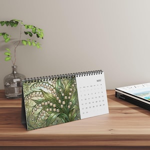 May include: A desk calendar with a black spiral binding featuring a May page. The left side displays a detailed illustration of white lily of the valley flowers with green leaves in a stained glass style. The right side shows the calendar grid for May.