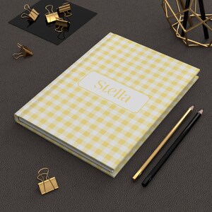 May include: A yellow and white gingham pattern journal with the name "Stella" in gold lettering. Gold and black pencils and gold binder clips are also on the dark gray surface.