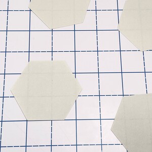 K&ouml;nnte beinhalten: Vier hellbeige sechseckige Teile sind auf einer wei&szlig;en Oberfl&auml;che mit einem blauen Gittermuster angeordnet. Die Teile scheinen aus Papier oder Karton zu bestehen und haben feine Gitterlinien aufgedruckt.