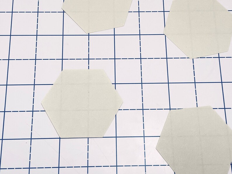 K&ouml;nnte beinhalten: Vier hellbeige sechseckige Teile sind auf einer wei&szlig;en Oberfl&auml;che mit einem blauen Gittermuster angeordnet. Die Teile scheinen aus Papier oder Karton zu bestehen und haben feine Gitterlinien aufgedruckt.