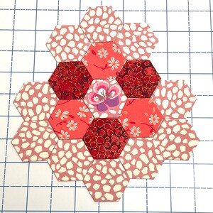 May include: A floral patchwork design made from hexagonal fabric pieces. The design includes floral, speckled, and deep red patterns. The central hexagon features a purple and white flower. The design is on a grid.