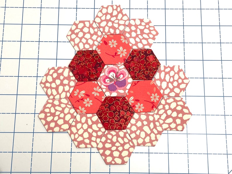 May include: A floral patchwork design made from hexagonal fabric pieces. The design includes floral, speckled, and deep red patterns. The central hexagon features a purple and white flower. The design is on a grid.