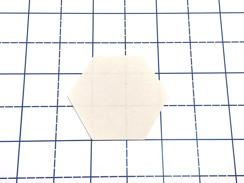 K&ouml;nnte beinhalten: Eine leere, cremefarbene, sechseckige Papierform, die sich auf einer wei&szlig;en Oberfl&auml;che mit einem blauen Rastermuster befindet. Das Raster besteht aus durchgezogenen und gestrichelten Linien.