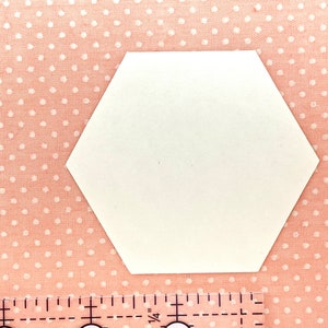 K&ouml;nnte beinhalten: Eine wei&szlig;e, sechseckige Papierform auf einem rosa Stoffhintergrund mit Polka Dots. Ein Lineal mit Zollmarkierungen ist unten im Bild zu sehen. Die Form ist wahrscheinlich f&uuml;r Bastelarbeiten oder zum Quilten gedacht.
