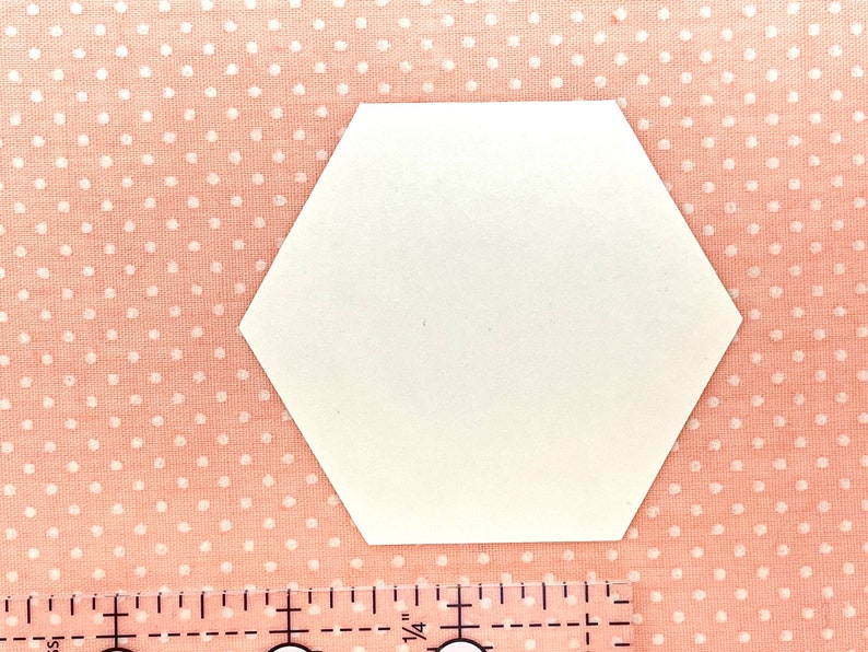 K&ouml;nnte beinhalten: Eine wei&szlig;e, sechseckige Papierform auf einem rosa Stoffhintergrund mit Polka Dots. Ein Lineal mit Zollmarkierungen ist unten im Bild zu sehen. Die Form ist wahrscheinlich f&uuml;r Bastelarbeiten oder zum Quilten gedacht.