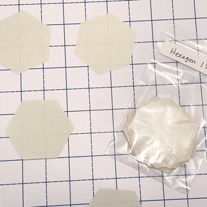 K&ouml;nnte beinhalten: Auf einer karierten Oberfl&auml;che liegen mehrere cremefarbene sechseckige Formen, jeweils etwa 3,8 cm breit. Ein klarer Beutel enth&auml;lt weitere Formen. Ein Etikett tr&auml;gt die Aufschrift "Hexagon 1 1/2 inch".